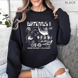 May include: Black crewneck sweatshirt with a white graphic design featuring the text "ARTEMIS II" and a detailed illustration of a space mission, including the NASA logo and the phrase "AROUND THE MOON FOR ALL HUMANITY."