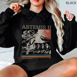 May include: Black hooded sweatshirt featuring a graphic design with the text "ARTEMIS II" and an illustration of astronauts, a planet, and space-related elements. The design includes the text "AROUND THE MOON" and the year "2026".
