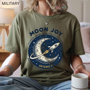 May include: Olive green t-shirt featuring a circular graphic with a crescent moon, rocket, and stars. The text reads "MOON JOY" and "ARTEMIS II", celebrating humanity's return to the lunar frontier. The shirt has short sleeves.