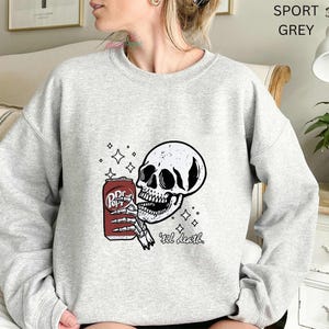 May include: Sport grey sweatshirt with a white skull graphic holding a Dr. Pepper can. The design includes the text "'Til death." and star shapes. The sweatshirt is a casual, comfortable style.