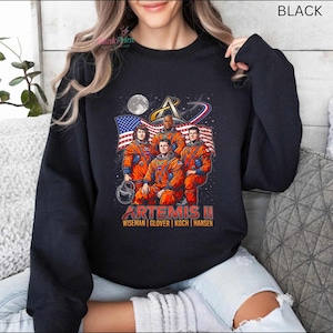 May include: Black crewneck sweatshirt featuring a graphic of the Artemis II mission crew in orange spacesuits, with the American flag and a moon in the background. The text "ARTEMIS II" and crew names are printed below the image.