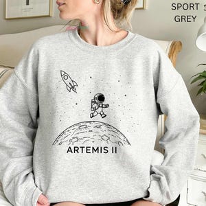 May include: Sport grey sweatshirt featuring a black and white graphic of an astronaut running on a moon with the text "ARTEMIS II". A rocket ship is also depicted in the design. The sweatshirt is a crew neck style.
