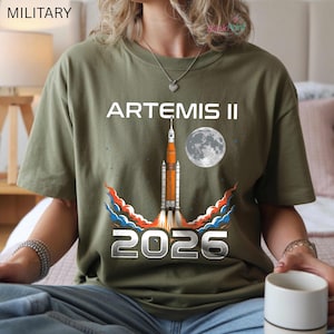 May include: Olive green t-shirt featuring the text "ARTEMIS II" above a rocket launching into space, with a full moon graphic and the year "2026" below. The rocket has orange and white details, with blue, orange, and red flames.