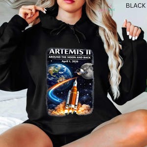 May include: Black hoodie featuring a graphic of the Artemis II mission. The design includes Earth, the Moon, a rocket, and the text "ARTEMIS II AROUND THE MOON AND BACK April 1, 2026". The hoodie is a casual garment.
