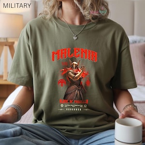 May include: Olive green t-shirt featuring the word "MALENIA" in red, above a graphic of a character with a golden helmet and red hair. Below the graphic are the words "BLADE OF MIQUELLA" and "UNBROKEN".