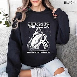 May include: Black sweatshirt with a white graphic that reads "RETURN TO THE MOON" above a triangular design featuring the moon and a stylized "A". The text "LUNAR FLYBY MISSION" is below the graphic.