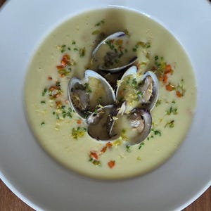 Puede incluir: Vista superior de un cuenco blanco lleno de sopa cremosa amarilla. Varias almejas abiertas están en el centro, adornadas con hierbas verdes picadas y pequeños trozos de ingredientes rojos y naranjas. El cuenco está sobre una superficie de madera.