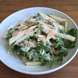 May include: A fresh salad on a white plate, featuring green leafy greens, sliced apples, and toasted almond slivers. The salad is lightly dressed and sprinkled with a white topping, likely cheese. The dish is set on a wooden table.