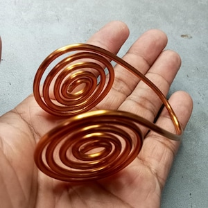 May include: Copper-toned arm cuff with two spiral motifs joined by a curved band. The cuff, crafted from a shiny metal, is designed for the upper arm. The spirals are the central design element, creating a striking visual effect.