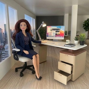 May include: A doll with long brown hair wearing a navy blue blazer and skirt sits in a white office chair at a desk. The desk features a computer monitor, a desk lamp, a small potted plant, a mug, and a notebook. A beige filing cabinet with three drawers sits under the desk.