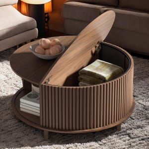 May include: A round, wooden coffee table with a lift-up lid revealing storage space. The table has a dark brown finish with vertical wooden slats around the base. A bowl of peaches sits on the tabletop, and a folded blanket is inside.