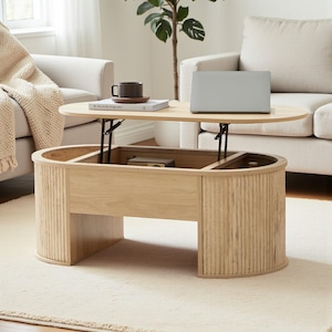 May include: A light wood oval coffee table with a lift-top design. The table has a lower storage area and is set on a light-colored rug. A laptop, a mug, and a book are on the table. The table is surrounded by light-colored furniture.