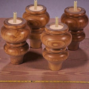 May include: Four wooden furniture legs with a turned design. Each leg is a warm brown color and features a threaded metal insert at the top. The legs are displayed on a wooden surface, showcasing their craftsmanship and potential for furniture projects.