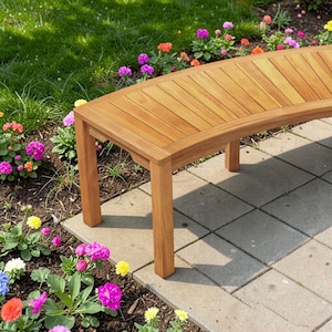 May include: A curved wooden bench with a slatted top and four square legs. The bench is made of light brown wood and sits on a gray stone patio. Colorful flowers and green grass surround the bench.