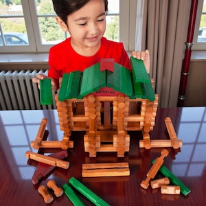 May include: A child is building a wooden log cabin toy. The cabin has a green roof and a red chimney. The words "LINCOLN LOGS" are printed on the roof. The toy is made of natural wood and green painted pieces.