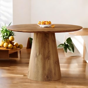 May include: A round wooden table with a light brown finish. The table has a single, conical base and a smooth tabletop. A small white bowl of oranges sits on the table. The table is surrounded by a light-colored wooden floor.
