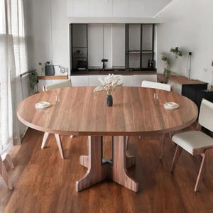 May include: A large, oval wooden dining table with a natural finish. The table has a central support and seating for several people. The table is surrounded by chairs and set with plates and glasses. The table is approximately 72 inches long.