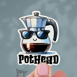 May include: A sticker featuring a cartoon illustration of a coffee maker wearing sunglasses, with the word "PotHead" in bold letters. The sticker has a white border and is held by a person's hand.