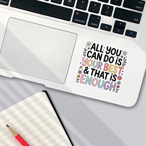 Puede incluir: Una pegatina blanca en un port&aacute;til con el texto "ALL YOU CAN DO IS YOUR BEST & THAT IS ENOUGH" en letras coloridas y detalles florales. Un cuaderno y un l&aacute;piz est&aacute;n en primer plano.
