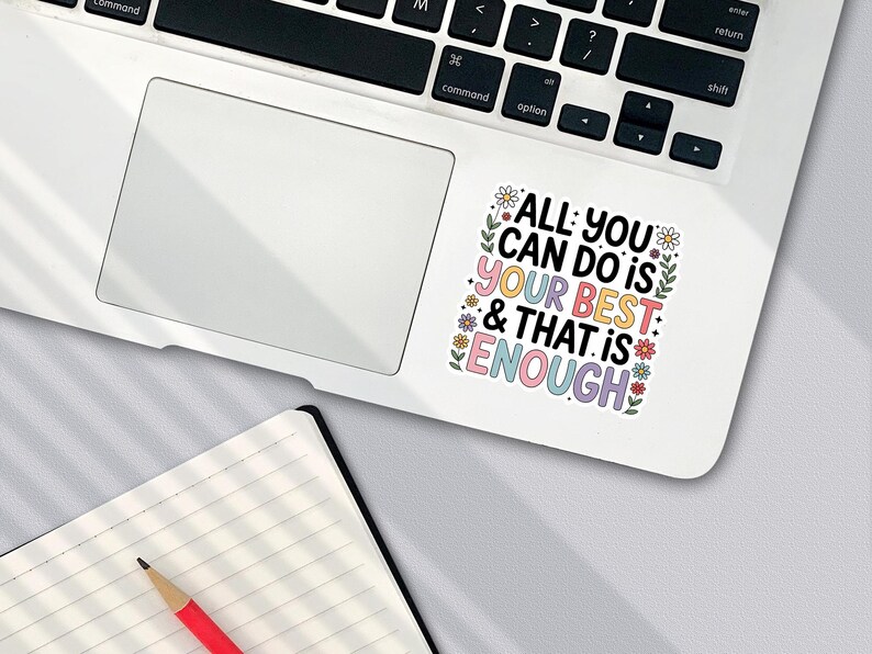 Puede incluir: Una pegatina blanca en un port&aacute;til con el texto "ALL YOU CAN DO IS YOUR BEST & THAT IS ENOUGH" en letras coloridas y detalles florales. Un cuaderno y un l&aacute;piz est&aacute;n en primer plano.