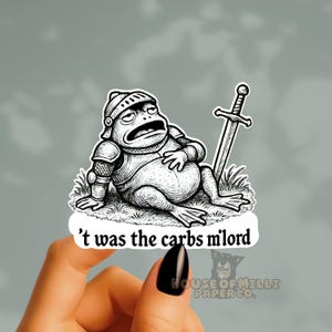 May include: Black and white sticker featuring a cartoon frog in medieval armor, resting on grass with a sword. The text "'t was the carbs milord" is below the frog. The sticker is held by a hand.