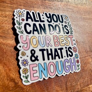 Puede incluir: Una pegatina cuadrada con el texto "ALL YOU CAN DO IS YOUR BEST & THAT IS ENOUGH" en negro, rosa, amarillo, azul y morado. La pegatina tiene un borde blanco con detalles florales y est&aacute; sobre una superficie de madera.