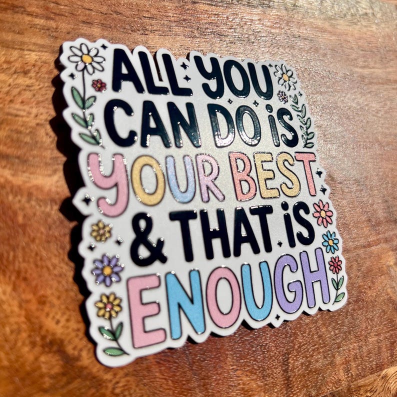 Puede incluir: Una pegatina cuadrada con el texto "ALL YOU CAN DO IS YOUR BEST & THAT IS ENOUGH" en negro, rosa, amarillo, azul y morado. La pegatina tiene un borde blanco con detalles florales y est&aacute; sobre una superficie de madera.