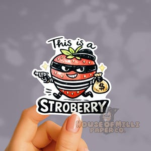Puede incluir: Una pegatina con una fresa de dibujos animados como un bandido, con una máscara negra, sombrero y camisa a rayas, sosteniendo una pistola y una bolsa de dinero. El texto dice "This is a" y "STROBERRY".