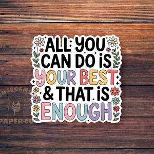 Puede incluir: Una pegatina colorida con el texto "ALL YOU CAN DO IS YOUR BEST & THAT IS ENOUGH". La pegatina tiene un borde blanco y est&aacute; decorada con flores y hojas. El fondo es una superficie de madera.