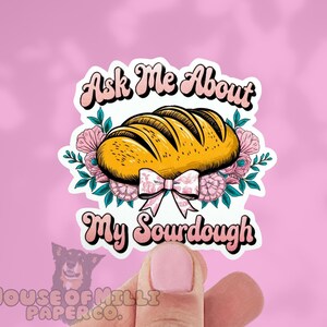 Puede incluir: Adhesivo con una ilustración de pan de masa madre, rodeado de flores rosas y un lazo. El texto "Ask Me About My Sourdough" está escrito en una fuente retro. Fondo rosa.