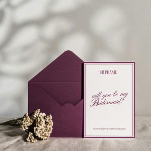 May include: A plum-colored envelope and invitation card set. The card reads "Stephanie, will you be my Bridesmaid?" in elegant script. A small bouquet of dried flowers rests near the stationery, perfect for wedding announcements.