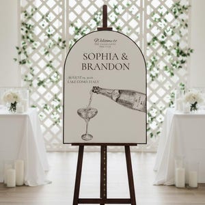 May include: A dark brown wooden easel displays a wedding welcome sign. The sign features the names "Sophia & Brandon", the date, and location. A champagne bottle pours into a glass, all in a black and white illustration.