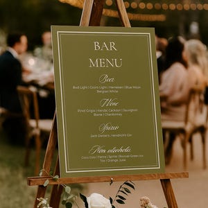 Könnte beinhalten: Ein hölzernes Staffelei zeigt eine grüne Barkarte mit weißem Text. Die Karte listet Bier, Wein, Spirituosen und alkoholfreie Getränke auf. Die Staffelei ist mit weißen und rosa Blumen geschmückt, mit einem verschwommenen Hintergrund.