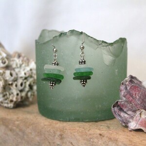 May include: Earrings made of layered sea glass in shades of green and blue-green, with silver-colored metal accents. The earrings are displayed against a blurred background with a green glass container and seashells.