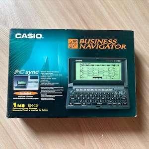 Puede incluir: Una caja de producto rectangular de color verde azulado oscuro para un organizador electrónico Casio Business Navigator BN-10. La caja muestra un gráfico del dispositivo con un teclado QWERTY y una pantalla monocromática verde con calendario.