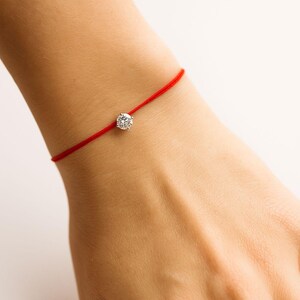 May include: A red string bracelet featuring a single, clear, round gemstone. The bracelet is simple and elegant, with the gemstone centered on the red string. The background is plain white.