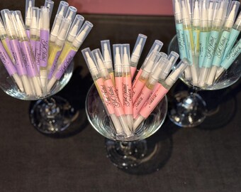 May include: An overhead shot displays three glass bowls filled with cuticle oil pens. The pens have clear tubes with white applicators and colored labels in pink, purple, and teal. The labels read "Get Nailed! Cuticle Oil."