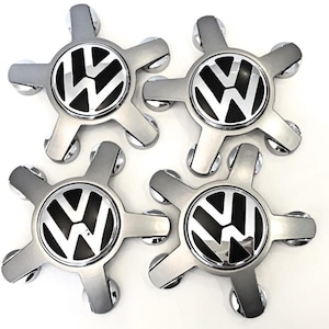 May include: Set of four silver-colored hubcaps with a black and white Volkswagen logo in the center. Each hubcap has a five-point star design with rounded edges, suitable for car enthusiasts.