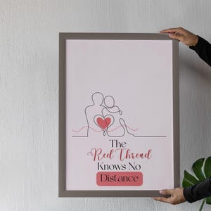 Pode incluir: Uma impressão emoldurada com um desenho de linha minimalista de duas figuras abraçando-se, com um coração no centro. O texto "The Red Thread Knows No Distance" é exibido em uma fonte cursiva, com um fundo retangular rosa.
