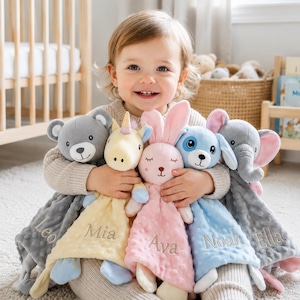 Puede incluir: Una colección de cinco mantitas de seguridad de peluche con cabezas de animales: un oso gris, un unicornio amarillo, un conejo rosa, un perro azul y un elefante gris. Cada manta suave con relieve está bordada con los nombres Leo, Mia, Ava, Noah y Ella.