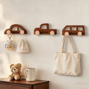 May include: Three wooden car-shaped wall hooks in varying designs, each with a light brown finish and light pink wheels. The name "Lucas" is inscribed on each hook. One hook holds a baby toy, another a small bag, and the third a tote bag.