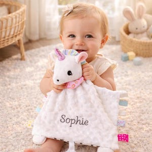 Könnte beinhalten: Eine weiße Einhorn-Kuscheldecke mit dem Namen "Sophie" aufgestickt. Das Einhorn hat ein rosa Horn und einen rosa und gemusterten Hals. Die Decke hat bunte Etiketten an den Rändern.