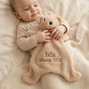 Könnte beinhalten: Ein beigefarbenes Teddybär-Kuscheltuch mit dem gestickten Text "Isla Mama 2026". Das Plüschtier hat eine weiche, strukturierte Oberfläche und eine schwarze Nase. Das Kuscheltuch ist für Babys konzipiert.