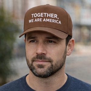 May include: A brown baseball cap with the text "TOGETHER, WE ARE AMERICA" in white. The cap has a curved brim and is worn by a person. The cap is a solid brown color.