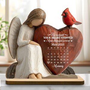 May include: A wooden angel figurine with a heart-shaped memorial. The heart is engraved with text and a calendar for May 2025. A red cardinal sits atop the heart. The angel is wearing a white dress and has wings.