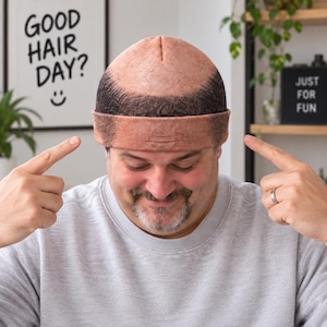 Peut inclure: Un bonnet en tricot arborant un motif imprimé réaliste représentant un crâne dégarni avec une peau claire sur le dessus et des cheveux brun foncé sur les côtés. Le bonnet présente un revers replié et une texture en tissu uniforme.