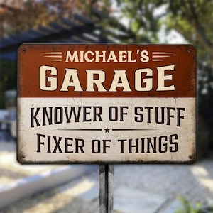 May include: A rectangular metal sign with a distressed finish featuring a brown top section and a white bottom section. The text reads MICHAEL'S GARAGE, KNOWER OF STUFF, and FIXER OF THINGS in bold, dark brown lettering with decorative lines and a star.