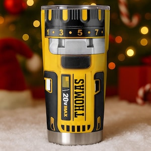 May include: A stainless steel tumbler with a yellow and black design resembling a power tool. The tumbler features the name "THOMAS" and "20V MAX" printed on it. The top has numbers 1, 3, 5, and 7. The background is blurred with Christmas decorations.