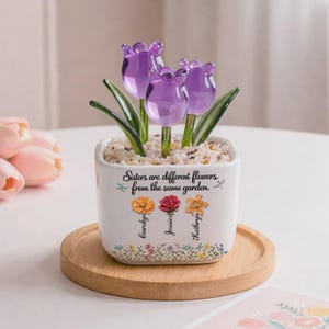 May include: A white ceramic planter with three purple glass tulip flowers. The planter has the text "Sisters are different flowers from the same garden" and the names Carolyn, Jessica, and Kathryn. The planter sits on a wooden tray.