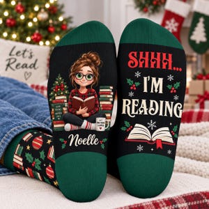 Può includere: Un paio di calzini neri e verde scuro con un personaggio dei cartoni animati che legge un libro, pile di libri, un albero di Natale e una tazza. Il testo recita SHHH... I'M READING e il nome Noelle, con motivi di agrifoglio e fiocchi di neve.